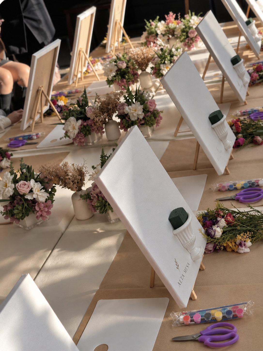 Estaciones de taller creativo con lienzos, flores, pinturas y herramientas para cada asistente
